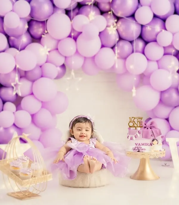 A perfect first birthday portrait. The birthday girl sits proudly in her purple dress, surrounded by a stunning balloon arch, a custom cake, and light-up letters spelling her initials.
