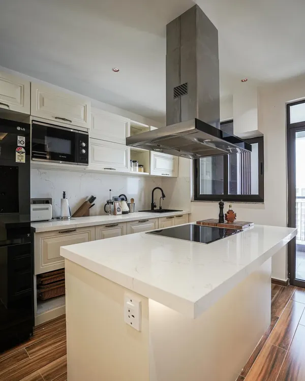 A modern kitchen with off-white shaker cabinets, a quartz island with a built-in cooktop, and a striking stainless steel chimney.