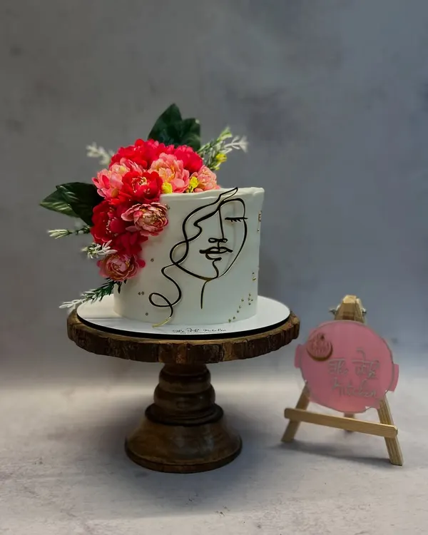 This cake features a minimalist one-line art face, hand-piped onto smooth white buttercream. The design is complemented by a vibrant bouquet of fresh flowers.