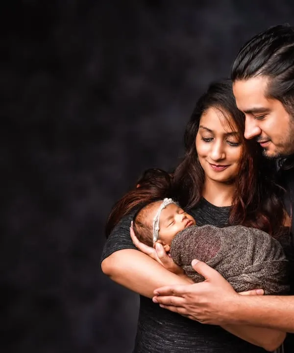A classic newborn portrait with parents. The dark, moody background creates an intimate atmosphere, drawing attention to the parents' gentle hold and loving gaze upon their new baby.