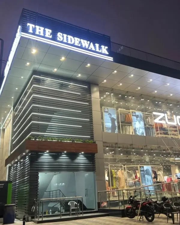 A night view of the completed commercial building, 'The Sidewalk'. The facade features horizontal louvers with integrated LED lighting, creating a clean, modern, and inviting look for the retail spaces within.