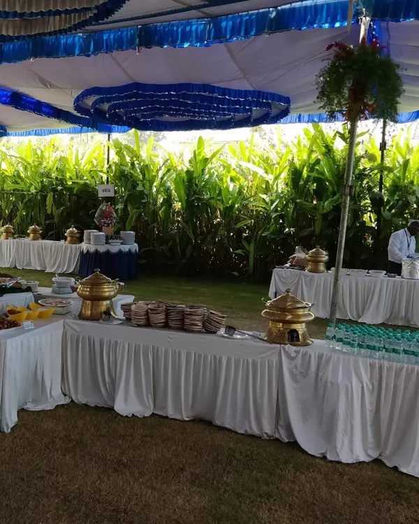 A beautiful outdoor catering setup under a tent for a day event. The multi-level buffet arrangement allows for a grand display of food against a lush green backdrop, perfect for weddings, receptions, and society events.