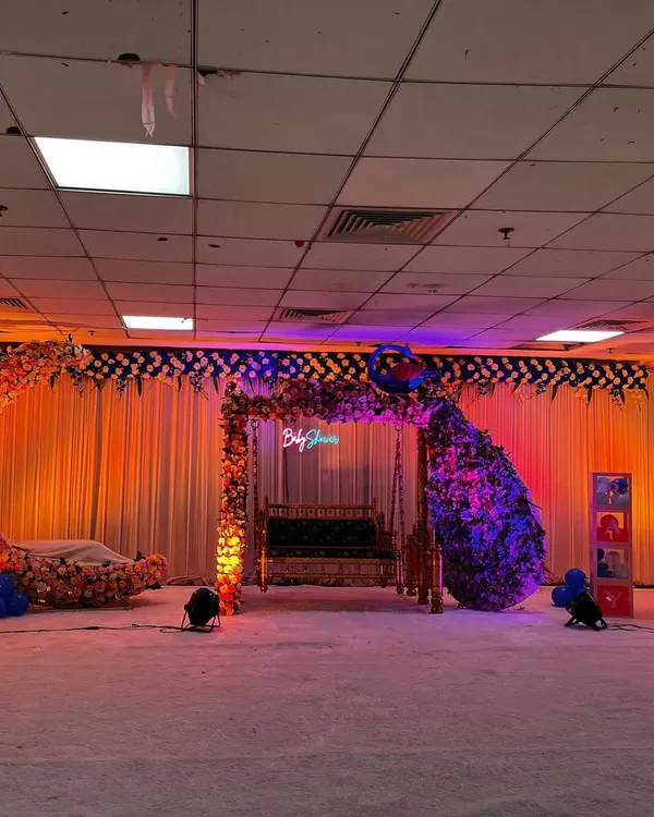 A beautiful stage setup for a baby shower featuring a floral moon-gate, a decorated Sankheda jhula, and a peacock made of flowers. The warm lighting creates a welcoming atmosphere for the celebration.