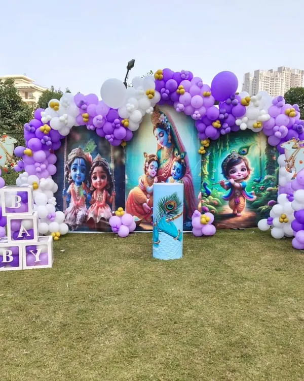 A unique Krishna-themed baby shower. This outdoor setup features a custom backdrop with adorable illustrations of baby Krishna, framed by a purple and white balloon arch.