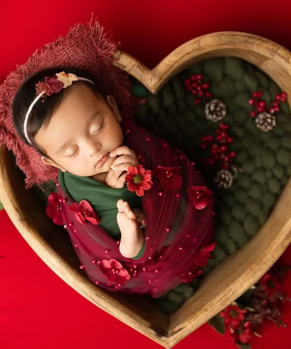 A wide shot of our Christmas-themed setup, with a peacefully sleeping newborn nestled in a heart-shaped bowl. The rich red backdrop and festive details create a warm, celebratory feel for your baby's first holiday photos.