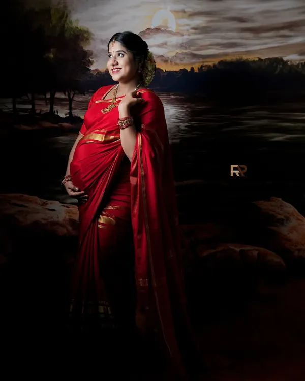 A portrait of timeless grace. The mom-to-be is dressed in a vibrant red saree, standing against a painted backdrop that evokes a serene, moonlit landscape. This is one of our signature traditional looks.