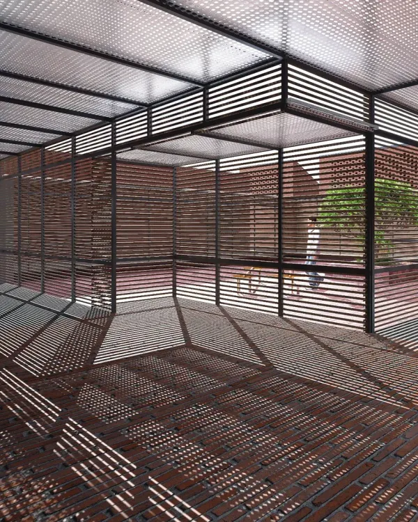 At Studio Tarai in Rudrapur, a louvered steel canopy casts a complex grid of shadows on the brick floor, creating a dynamic, patterned space that changes with the movement of the sun.