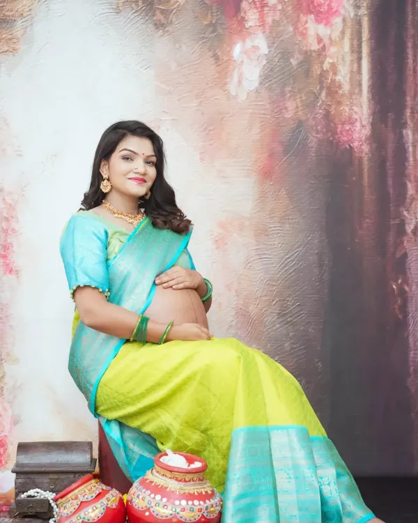 A graceful solo portrait of an expecting mother in a traditional silk saree. The painterly backdrop and classic props like decorated pots add a rich, cultural feel to this maternity photo.
