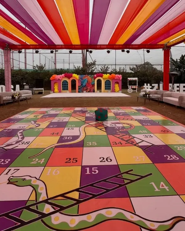 A wide shot of the pre-wedding game night setup. Under a colorful draped ceiling, the life-sized Snakes and Ladders board takes center stage, promising a fun and interactive experience for everyone.