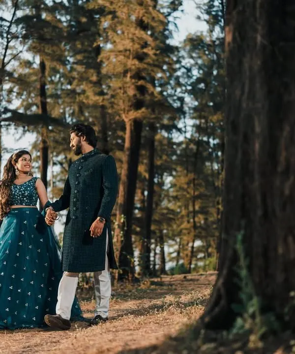 A couple posing in a beautiful forest setting. He wears a classic dark sherwani that perfectly complements her teal lehenga, showcasing my coordinated rental options.