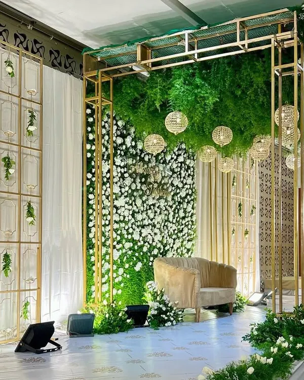 A wide view of this spectacular reception stage. The design features a lush green floral ceiling, a white flower wall, and elegant gold-toned structural elements for a modern and glamorous look.
