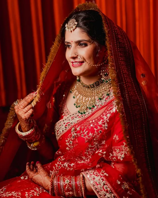 A classic portrait of a North Indian bride in her radiant red lehenga. The way she adjusts her ghunghat (veil) is a timeless pose that showcases both her traditional attire and her joyful anticipation.