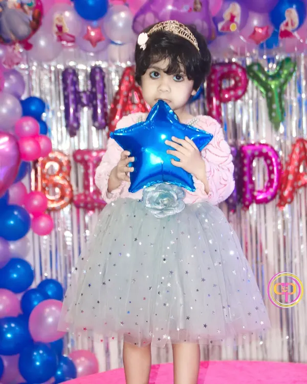 A young birthday girl dressed as a princess, posing in front of her colorful balloon and silver foil curtain backdrop.