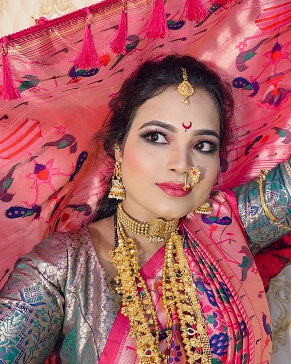 A classic Maharashtrian bride, holding the veil. The look features gold shimmery eyeshadow, winged liner, and a traditional `nath` that perfectly complements the pink Paithani saree.