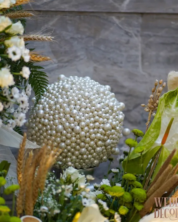 Every detail whispers elegance. Here, a sphere covered entirely in pearls sits within a lush arrangement of white and green florals, showcasing our commitment to creating unique, luxurious textures.