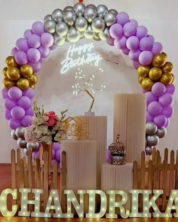 This elegant setup is perfect for milestone birthdays. It features a purple, gold, and silver balloon arch, my 'Happy Birthday' neon sign, and custom light-up marquee letters, all framed by a small picket fence for a finished look.