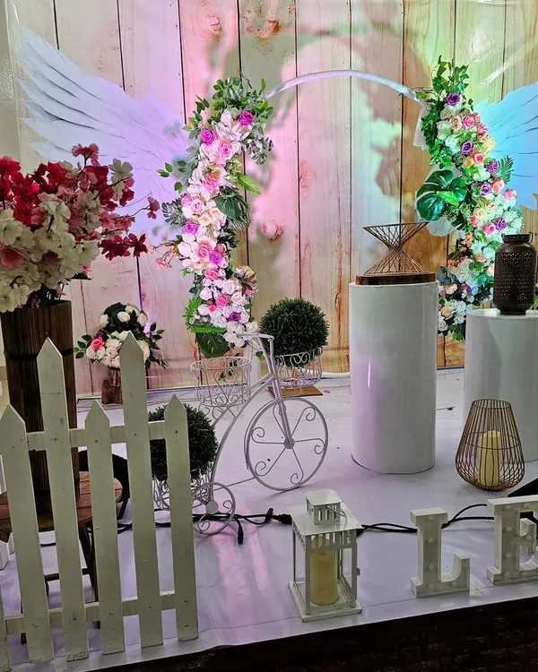 A whimsical and romantic backdrop featuring angel wings and fresh floral arches. The rustic wood-paneled background and white picket fence add to the garden-party charm.