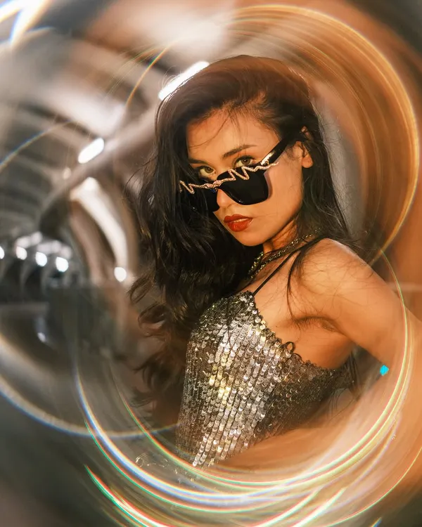 A stylish portrait of a woman in a sequined top and unique sunglasses, framed by swirling lights from a creative filter. The setting appears to be a modern, metallic tunnel, adding to the futuristic vibe.