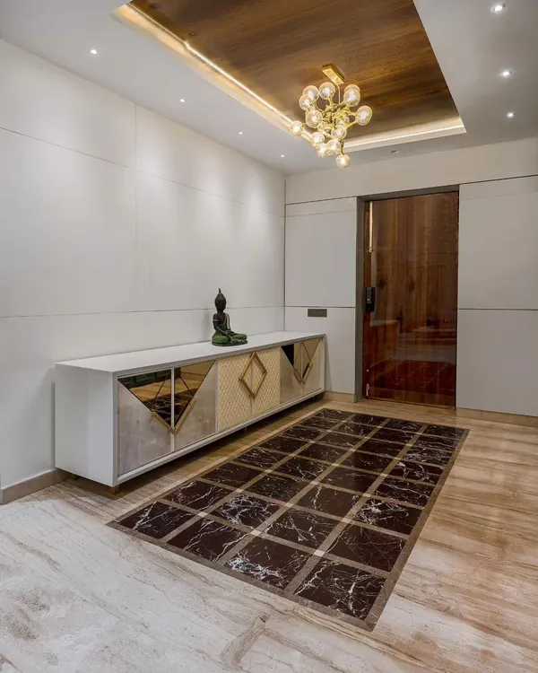 This contemporary foyer design creates a lasting first impression. A custom console with mirrored and geometric-patterned doors sits below a wooden coffered ceiling with a modern chandelier, while a marble floor inlay adds a touch of luxury.