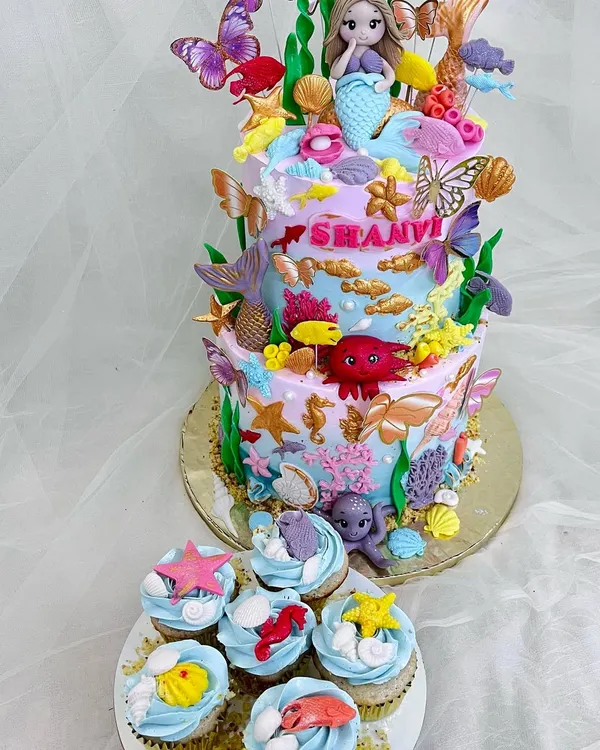 An enchanting two-tier mermaid cake surrounded by matching under-the-sea themed cupcakes. This creation is a deep dive into detail, with handcrafted fondant sea creatures, shimmering shells, and beautiful butterflies.