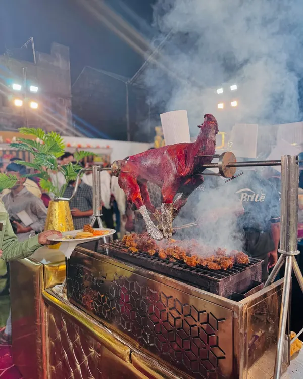 A whole tandoori goat roasting on a rotisserie, with smoke billowing from the grill. This is a spectacular centerpiece for any barbecue station.