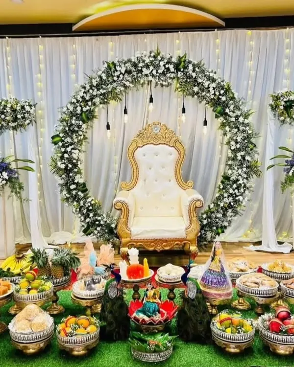 A beautiful Half-Saree ceremony stage with a white and green floral ring, a royal throne chair, and a traditional display of offerings.