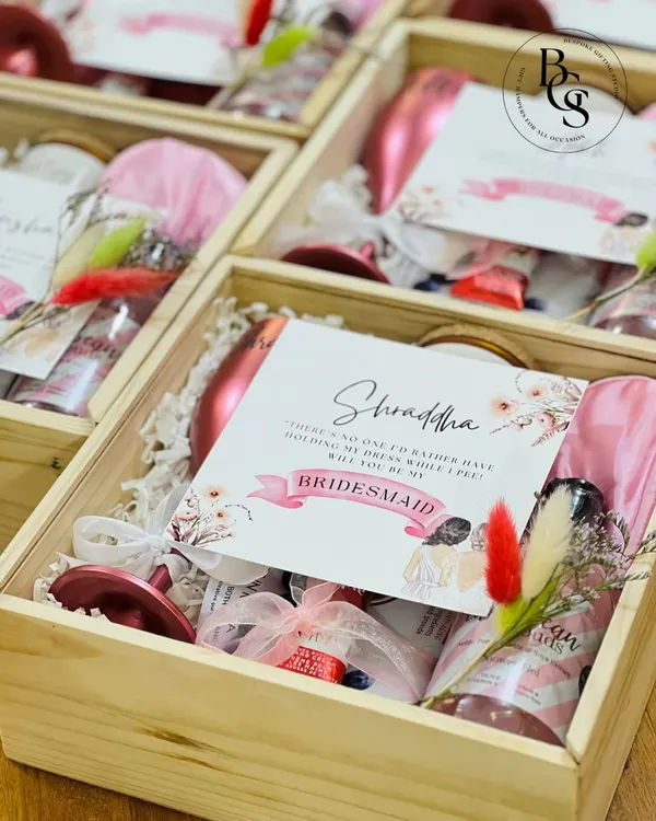 A personalized bridesmaid proposal hamper in a wooden crate. It's filled with thoughtful items like a custom wine glass, a bottle of sparkling rosé, a satin eye mask, and a card that perfectly pops the question.