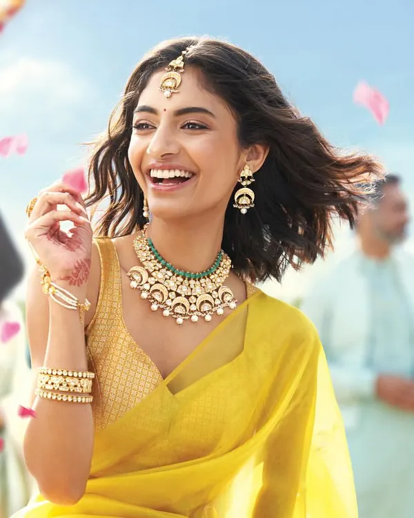 A candid, joyful moment from a Tanishq campaign, styling a modern Polki and emerald necklace set. The look is fresh and contemporary, pairing the jewellery with a simple yellow saree to appeal to a younger audience celebrating traditional events.