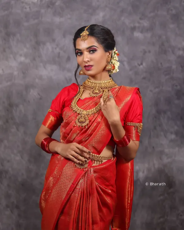 This studio shot captures the elegance of a classic red bridal saree. The focus is on the rich fabric, intricate gold jewelry, and the model's graceful pose, all against a simple, textured grey backdrop to make the colors pop.