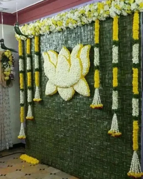 A beautiful Haldi backdrop made from a woven green mat, adorned with a lotus motif made of white petals and hanging garlands of yellow and white flowers.