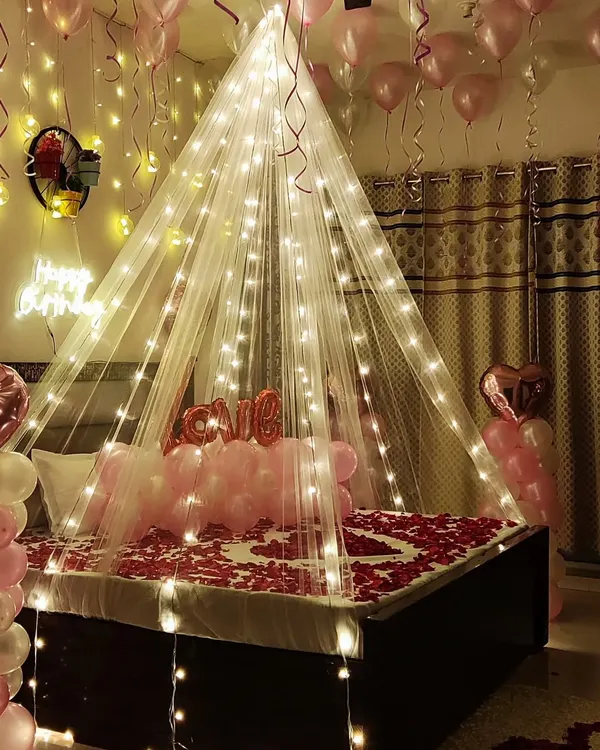 A dreamy birthday bedroom surprise featuring a sheer canopy illuminated with warm fairy lights. The decor includes soft pink and white balloons, rose petals arranged in a heart on the bed, and a "love" foil balloon to complete the romantic atmosphere.