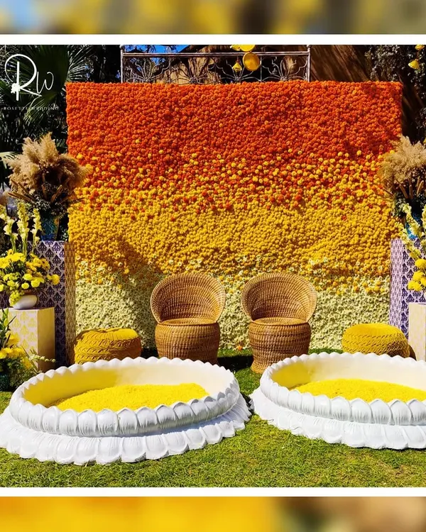 A stunning Haldi backdrop made entirely of flowers in shades of orange and yellow, creating a beautiful ombré effect. The seating is completed with wicker chairs and lotus-shaped basins for the ceremony.