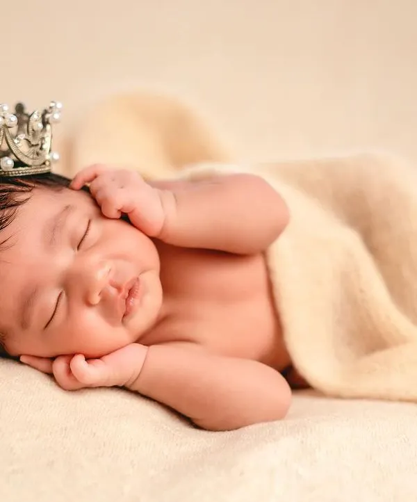 A classic "head on hands" pose on a soft, neutral blanket. This angle beautifully highlights the baby's delicate features and full head of hair, topped with a tiny crown.