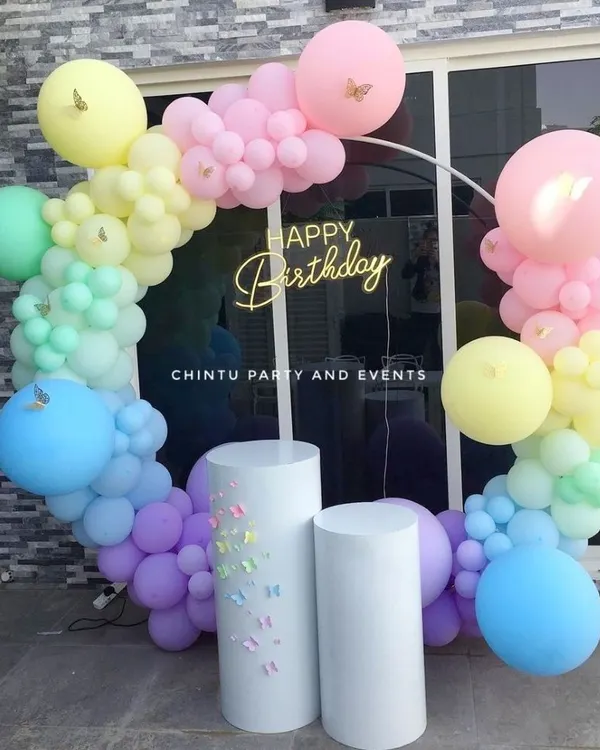 An outdoor birthday celebration featuring a pastel rainbow balloon ring. The setup is completed with a 'Happy Birthday' neon sign and white pedestals for the cake.