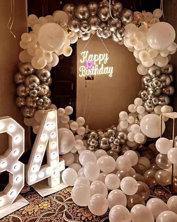 A glamorous 34th birthday setup featuring a circular balloon arch of white and chrome silver balloons, a 'Happy Birthday' neon sign, and large light-up numbers.