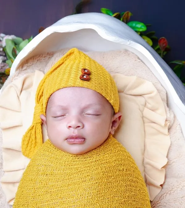 A close-up of a sleeping newborn in a yellow wrap and hat, nestled in a cozy prop. The focus is on the baby's peaceful face and tiny details.