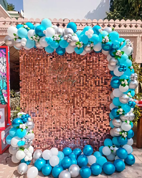 A breathtaking photo backdrop featuring a rose gold sequin wall. The wall is framed by a beautiful balloon arch in shades of turquoise, white, and silver, with delicate flower accents.