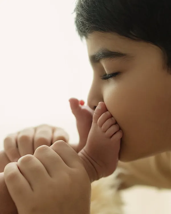 The beginning of a lifelong bond. An older brother gently kisses his newborn sibling's tiny foot, a moment of pure, unscripted love.
