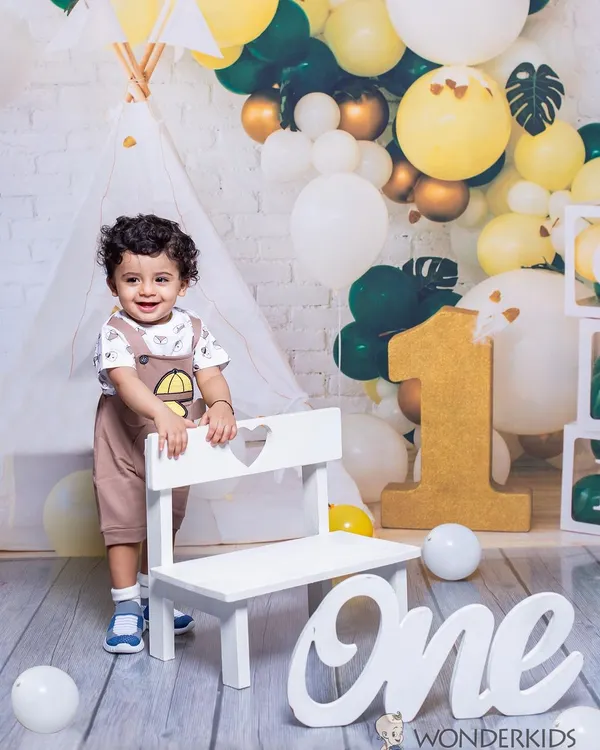 A happy toddler celebrating his first birthday. He stands proudly next to a "One" sign in a festive setup with a teepee and a beautiful balloon arch.