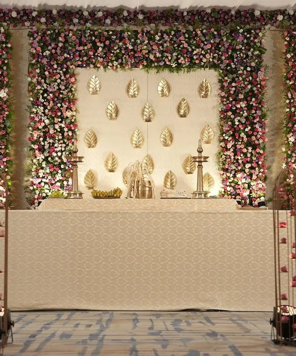A graceful cream and gold stage for a South Indian indoor wedding. The backdrop features gold leaf motifs, framed by a dense wall of pink and purple flowers, with an aisle lit by candles and hanging lamps.