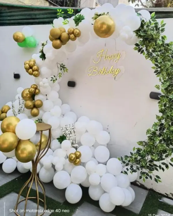 A beautiful and organic-style ring backdrop for an outdoor party. This design features a partial arch of white and gold balloons on one side and a cascade of artificial leaves on the other.