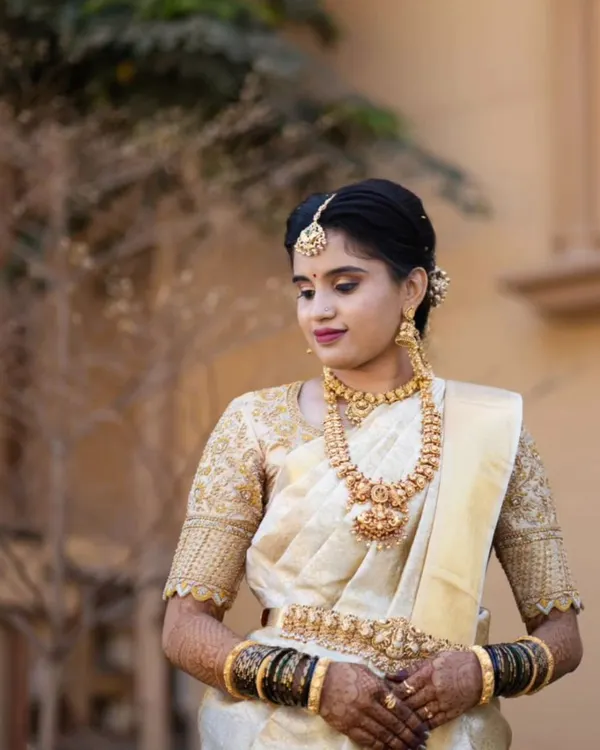 This bride looks serene and elegant for her muhurtham. Her makeup is classic and understated, with a focus on flawless skin and traditional gold jewellery that highlights her beautiful cream and gold silk saree.