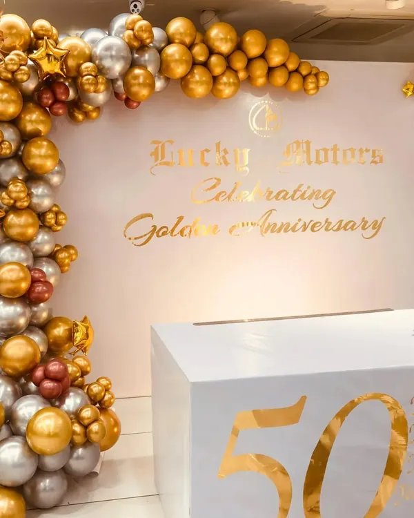 A clean and professional photo wall designed for the Lucky Motors 50th Anniversary. The chrome gold and silver balloon arch perfectly complements the gold vinyl lettering, creating a sophisticated branding moment for the corporate celebration.