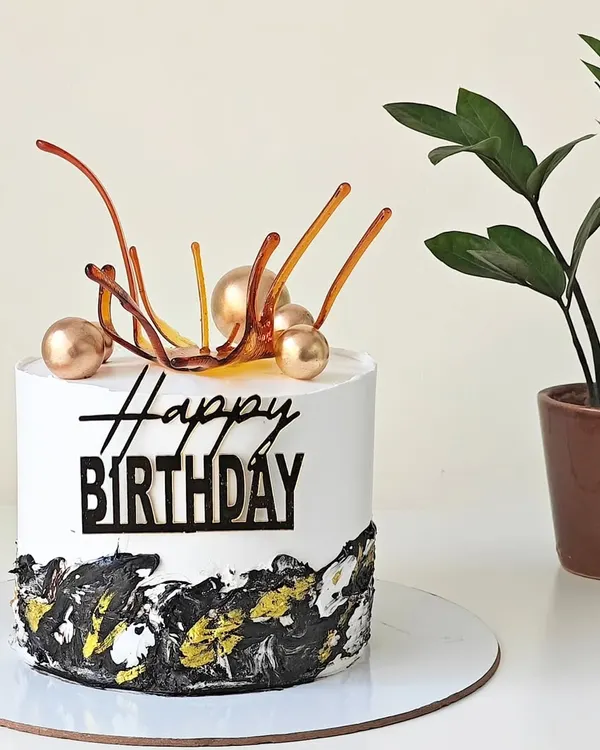 A modern birthday cake with a black, white, and gold abstract design at the base. The cake is topped with a pulled isomalt sugar sail and gold spheres for an artistic touch.
