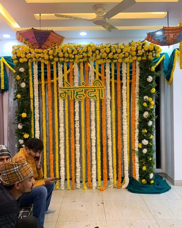 A classic Haldi and Mehndi backdrop featuring strings of fresh yellow and white flowers with a 'Mehndi' sign in Hindi. This setup is perfect for home functions and can be customized to fit your space.
