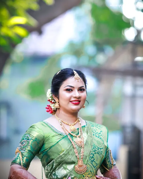 A radiant bridal portrait captured outdoors. The natural light beautifully highlights the rich green of her silk saree and the intricate gold jewelry.