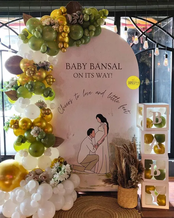 A beautiful, personalized baby shower backdrop for Baby Bansal. The custom illustration of the parents-to-be adds a deeply personal touch, complemented by an elegant olive green and gold balloon arch.
