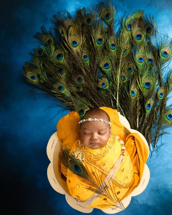 An overhead view of a newborn in a Krishna-themed setup, resting in a basket with a backdrop of fanned peacock feathers. The vibrant yellow wrap and blue background create a striking image.