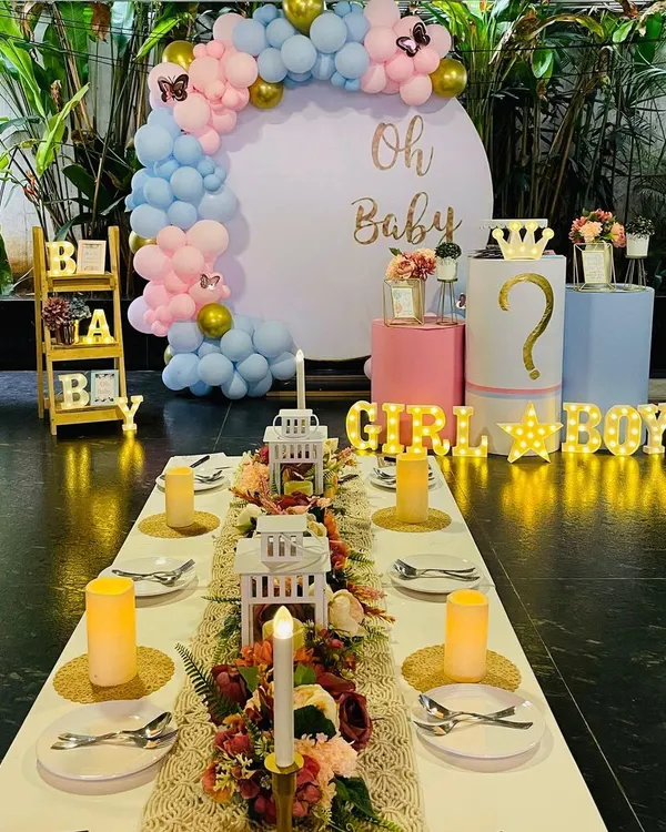 A beautiful baby shower setup combining a dining table arrangement with a photo backdrop. The 'Oh Baby' installation features a pastel balloon arch and marquee lights for the gender reveal.