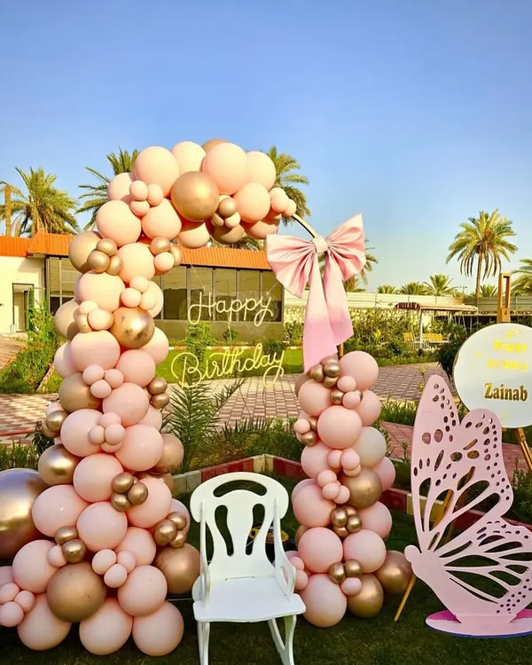 An outdoor butterfly-themed birthday setup with a beautiful pink and gold balloon arch. The decor includes a large pink butterfly prop, a 'Happy Birthday' neon sign, and a special chair for the birthday girl.